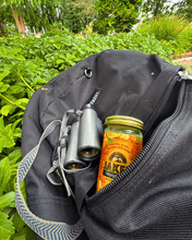 Load image into Gallery viewer, A backpack laying in the grass with a 4oz bottle of Jake's in its pouch.