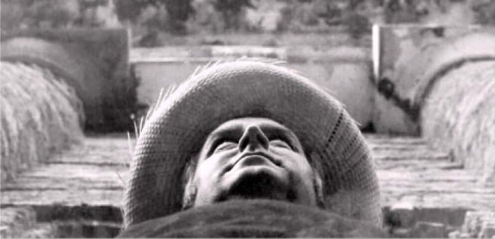 Portrait of Jake in his black shirt and straw hat beneath a stone arch. 